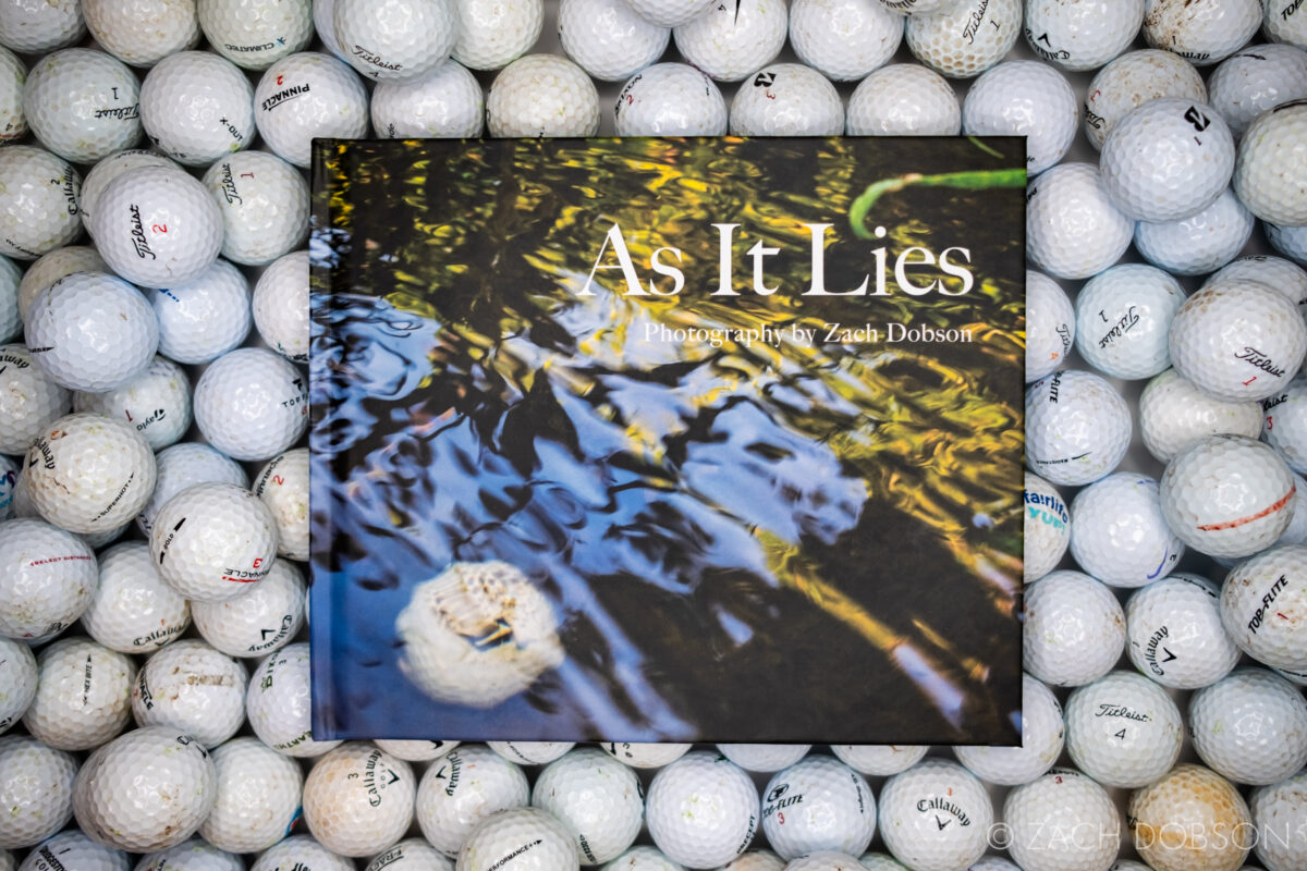 Cover photo of the As It Lies book laying on top of recovered golf balls in a bin