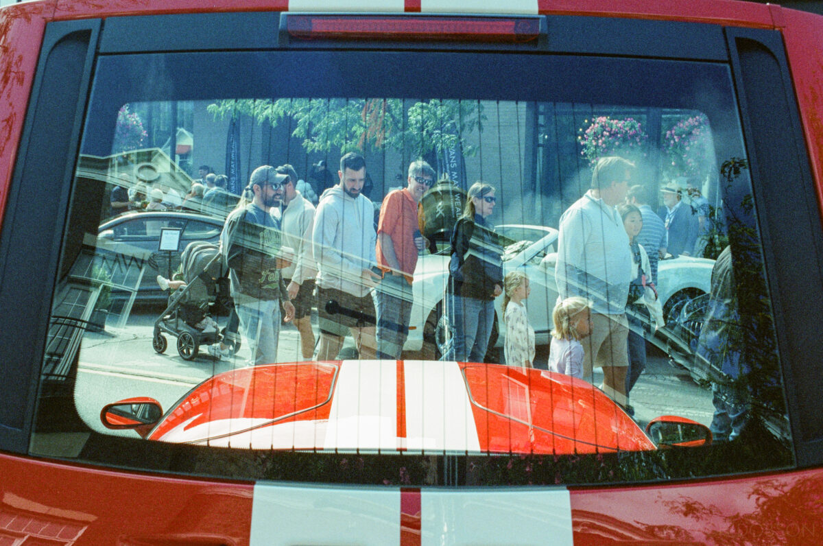 The rear hood of a Ford GT that covers its engine is open and the window frames the surrounding scene, but this time it's a film photo