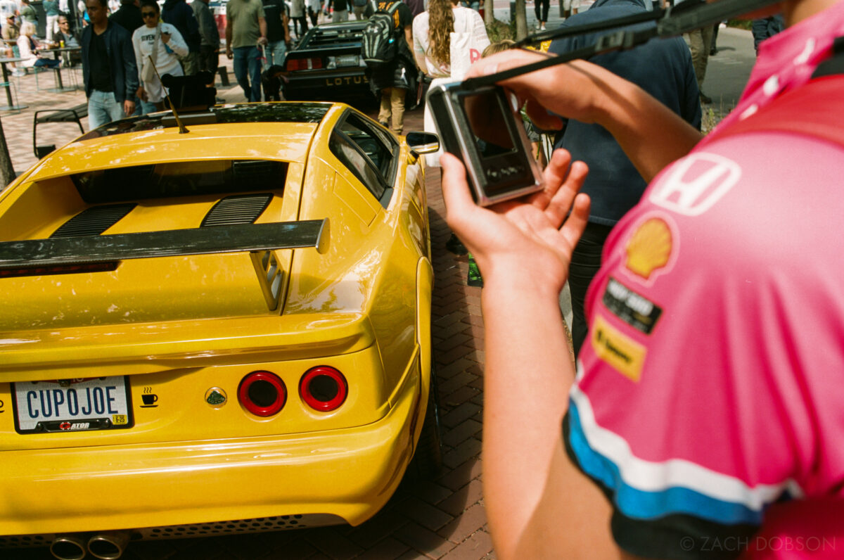 A person in a pink jersey photographs a bright yellow Lotus. Carmel Artomobilia 2024