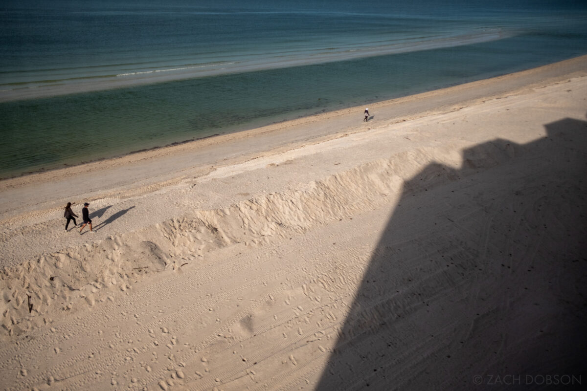 Building shadow and people walking along the beach with calm, dark blue waters. Bonita Springs, Florida.